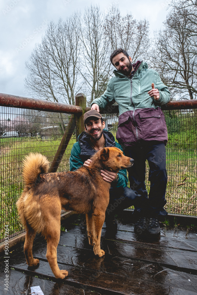 Two happy, young, caucasian and handsome brothers with jacket and backpacks petting two beautiful brown dogs in a wooden bridge in the forest in a rainy day, Valdivia, Chile Two happy, young, caucasian and handsome brothers with jacket and backpacks petting two beautiful brown dogs in a wooden bridge in the forest in a rainy day, Valdivia, Chile
