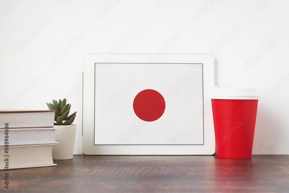 National flag of Japan on the tablet, textbooks, a red cup of hot drink coffee or tea on the table, the concept of learning Japanese National flag of Japan on the tablet, textbooks, a red cup of hot drink coffee or tea on the table, the concept of learning Japanese