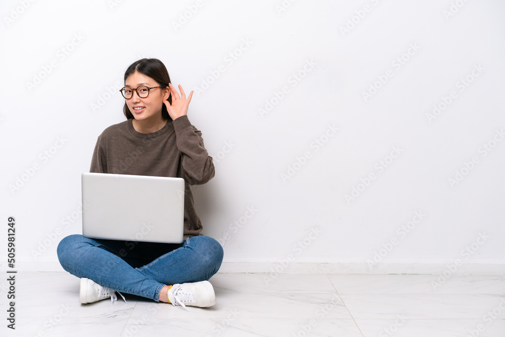 Young woman with a laptop sitting on the floor listening to something by putting hand on the ear Young woman with a laptop sitting on the floor listening to something by putting hand on the ear