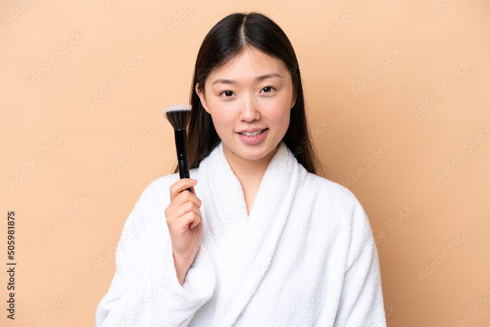 Young Chinese woman isolated on beige background holding makeup brush and whit happy expression Young Chinese woman isolated on beige background holding makeup brush and whit happy expression