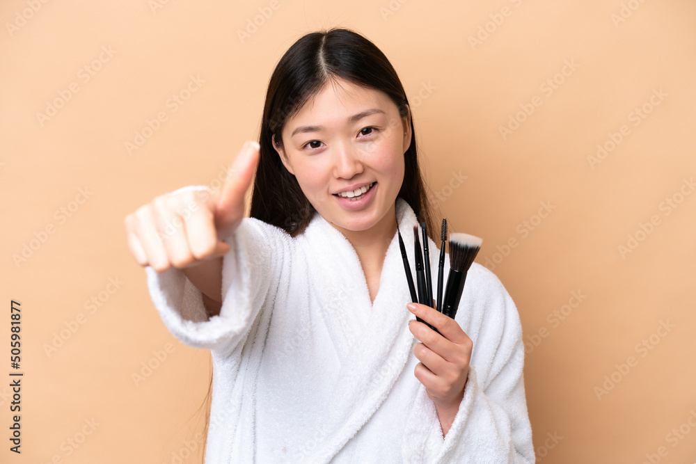 Young Chinese woman isolated on beige background holding makeup brush and with thumb up Young Chinese woman isolated on beige background holding makeup brush and with thumb up