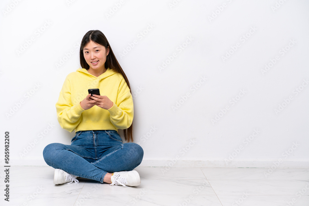 Young Chinese woman sitting on the floor isolated on white wall sending a message with the mobile Young Chinese woman sitting on the floor isolated on white wall sending a message with the mobile