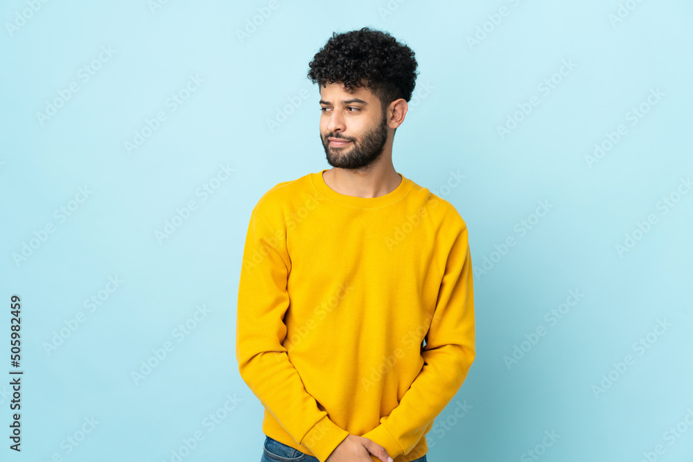 Young Moroccan man isolated on blue background having doubts while looking side Young Moroccan man isolated on blue background having doubts while looking side