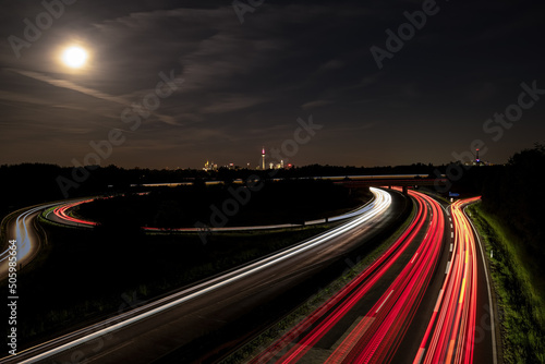 Die Skyline von Frankfurt am Main - im Vordergrund eine Autobahn mit Lichtspuren von Fahrzeugen bei Nacht und Mondlicht.