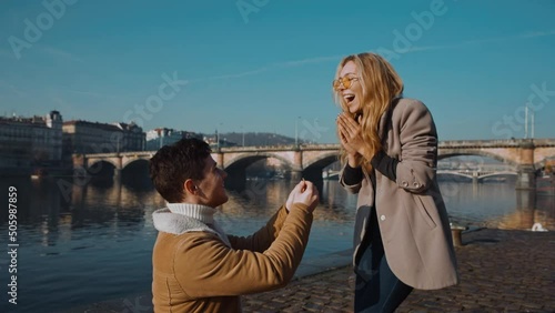 happy couple marriage proposal young man proposing to girlfriend with wedding ring enjoying surprise engagement. European town, pier, bridge on background photo