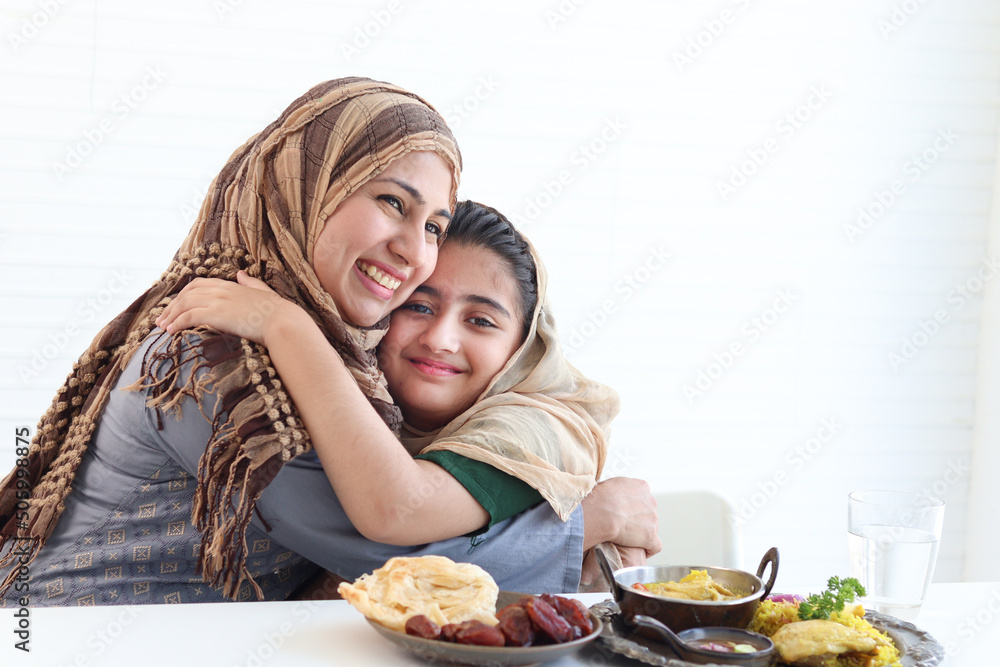 Φωτογραφία Adorable Pakistani Muslim girl sits at kitchen table with traditional Islamic halal food, kid with hijab hugs mother, warm love in family of mom and daughter, happy Islamic family on white background
