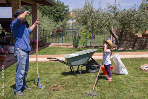 Photo of a gardener father and daughter wiping sweat from their foreheads as they rake and aerate the lawn at home.