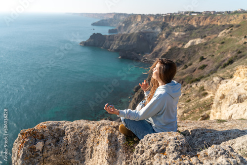Woman tourist enjoying the sunset over the sea mountain landscape. Sits outdoors on a rock above the sea. She is wearing jeans and a blue hoodie. Healthy lifestyle, harmony and meditation