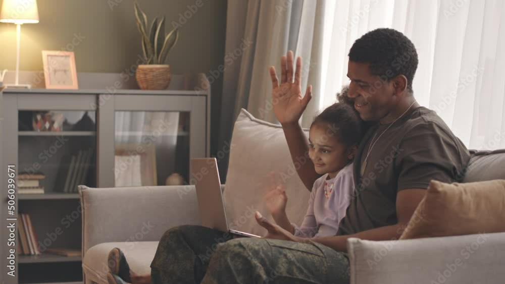 Medium slowmo of smiling Black man in military uniform and his 6 year old daughter waving at laptop camera while having video chat with mom off on assignment, sitting together on sofa at home Medium slowmo of smiling Black man in military uniform and his 6 year old daughter waving at laptop camera while having video chat with mom off on assignment, sitting together on sofa at home