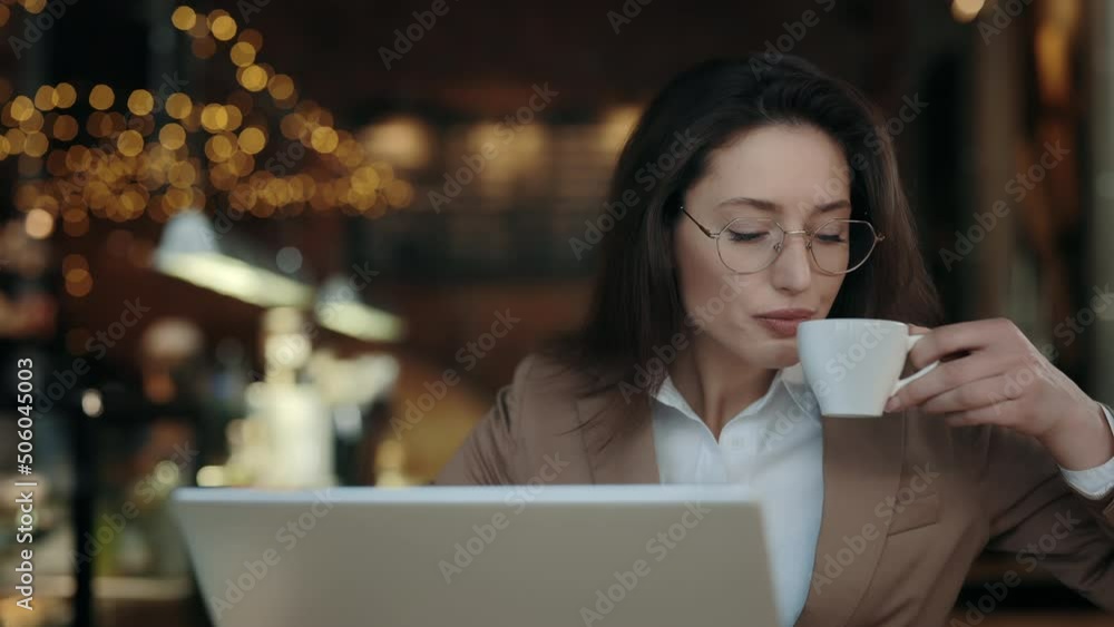 Charming business woman enjoying freshly brewed coffee while working on modern laptop at cafe. Caucasian lady with brown hair wearing eyeglasses and stylish formal jacket. Charming business woman enjoying freshly brewed coffee while working on modern laptop at cafe. Caucasian lady with brown hair wearing eyeglasses and stylish formal jacket.
