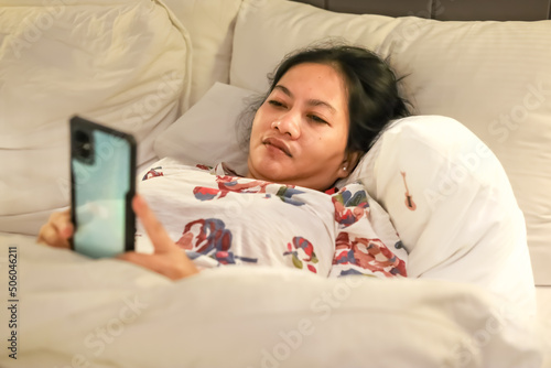 Smiling Asian woman wearing knitted sweater pink cold looking at mobile phone lying down on white bed. Beautiful girl typing on smartphone and surfing the net.Concept woman lifestyle and winter