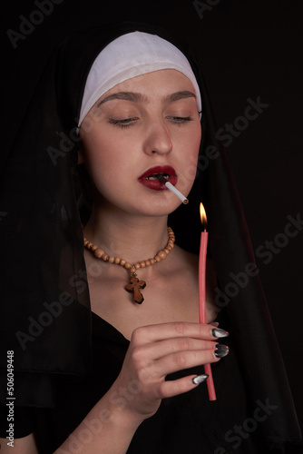 Portrait of a young nun lights up cigarette using a candle. Bad habits and blasphemy concept. Isolated on black background. photo
