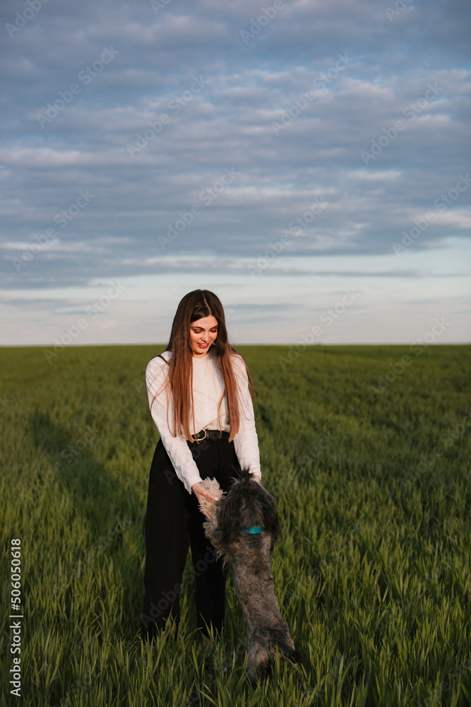 A stylish girl in a white shirt and black trousers with long hair plays with her dog in a field in nature at sunset.