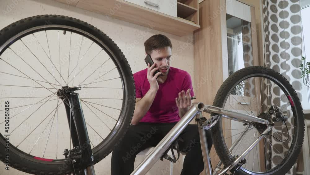 Upset man calls bicycle repair service on smartphone talks about broken bike. Cyclist sits near disassembled inverted bicycle with metal bottom bracket in hand in room. Guy is nervous, turns wheels. Upset man calls bicycle repair service on smartphone talks about broken bike. Cyclist sits near disassembled inverted bicycle with metal bottom bracket in hand in room. Guy is nervous, turns wheels.