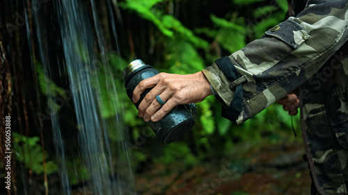 Hands of European young hiker hold glass, draws water from waterfall for drinking, in middle of rainforest