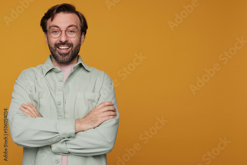 indoor portrait of handsome bearded male posing over orange background keeps his hands crossed on chest, looking into camera with shocked facial expression and smiles broadly