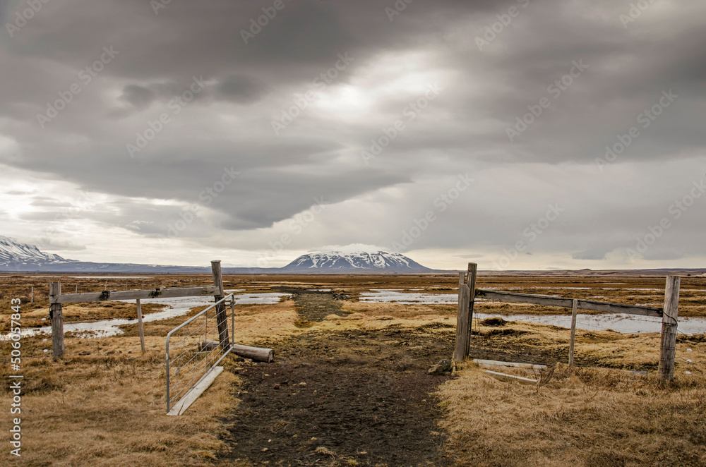 Haphazard wooden fence and iron gate in a watery landscape with shallow puddles and low vegetation and in the distance mount Sellandafjell under a dramatic sky Haphazard wooden fence and iron gate in a watery landscape with shallow puddles and low vegetation and in the distance mount Sellandafjell under a dramatic sky