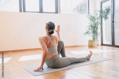 Back view of the senior woman practicing yoga, doing Half lord of the fishes exercise. Lady sitting in yoga posture and having morning meditation at home. Physical and spiritual practice concept