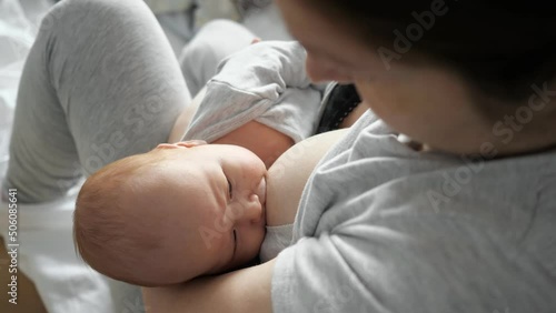 Mother feeds newborn baby with breast on bed at home. Adorable little girl in grey bodysuit sucks nipple lying in woman arms close upper view
