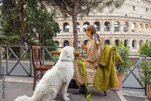 Photography Woman sitting with her dog at outdoor cafe near coliseum, the most famous landmark in Rome