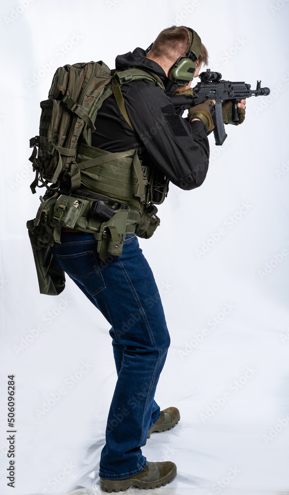 Fototapeta premium an airsoft soldier in full gear with his back to us is aiming at an optical sight. a man in an outfit, in headphones, a bulletproof vest, with a backpack and a belt. White background.