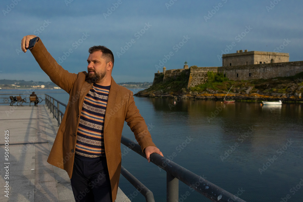 A man in a coat takes a selfie by the ocean.