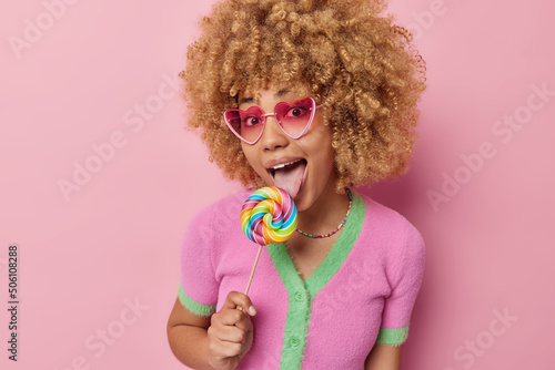 Cheerful curly haired woman licks delcious caramel multicolored round candy being sweet addicted wears trendy pink sunglasses casual shirt isolated over pink background. Sugar nutrition concept