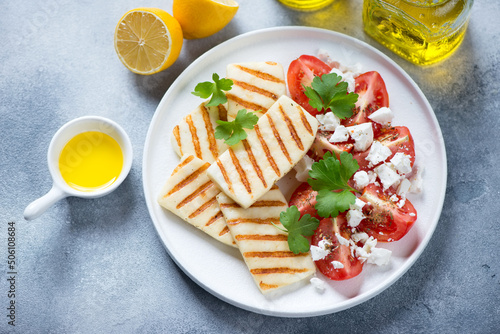 Grilled halloumi cheese with red tomatoes and feta on a white plate, high angle view on a light-blue stone background, horizontal shot