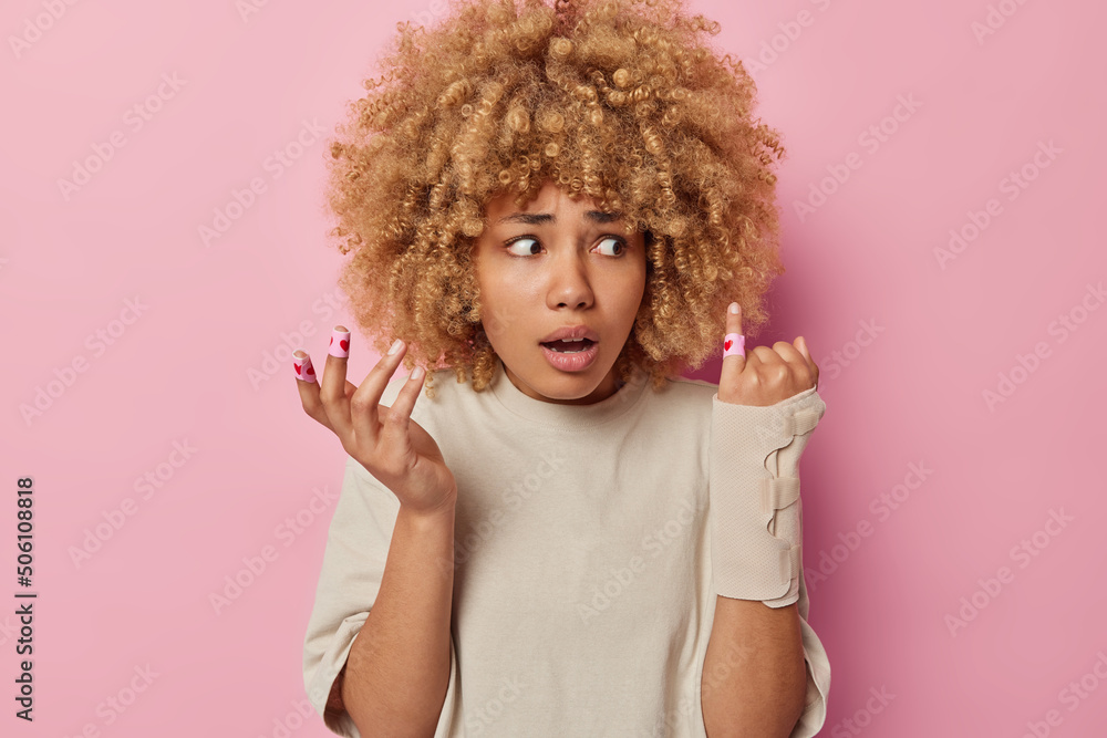 Worried curly haired woman has broken arm and injured finger wears bandage and plaster dressed in casual clothes isolated over pink studio background. Rehabilitation and health care concept.