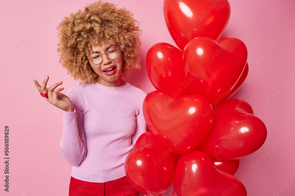 Dejected frustrated curly haired woman cries from despair looks doleful has spoiled makeup dressed in casual clothes celebrates lovers day alone poses near bunch of red balloons poses indoor Dejected frustrated curly haired woman cries from despair looks doleful has spoiled makeup dressed in casual clothes celebrates lovers day alone poses near bunch of red balloons poses indoor