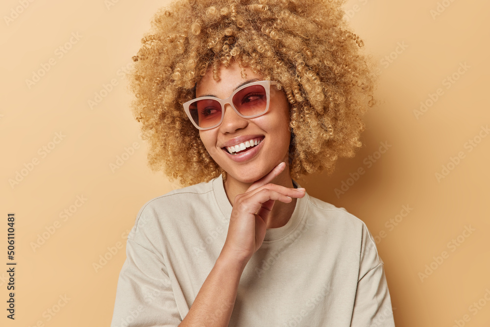 Happy curly haired woman touches jawline gently smiles broadly looks away wears sunglasses and casual t shirt feels glad dressed in casual t shirt isolated over brown background. People joy concept