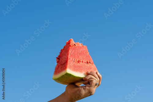 Male hand holding a slice of watermelon against a blue sky.