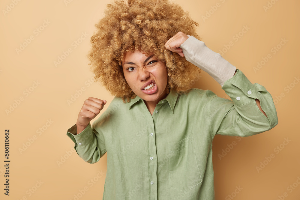 Outraged curly haired young woman clenches fists and looks angrily has broken arm wears bandage dressed in casual green shirt isolated over brown background. People aggression rage and anger Outraged curly haired young woman clenches fists and looks angrily has broken arm wears bandage dressed in casual green shirt isolated over brown background. People aggression rage and anger