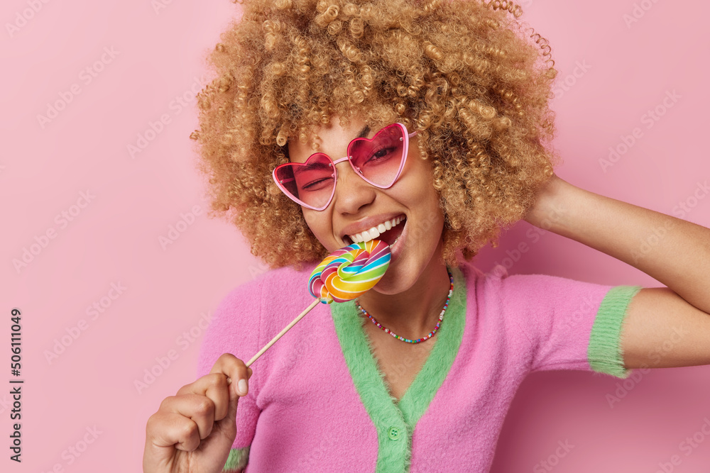 Happy carefree curly haired woman cannot live without sweets bites multicolred candy wears heart shaped sunglasses and t shirt isolated over pink background. Taste rainbow. Sweet life concept Happy carefree curly haired woman cannot live without sweets bites multicolred candy wears heart shaped sunglasses and t shirt isolated over pink background. Taste rainbow. Sweet life concept
