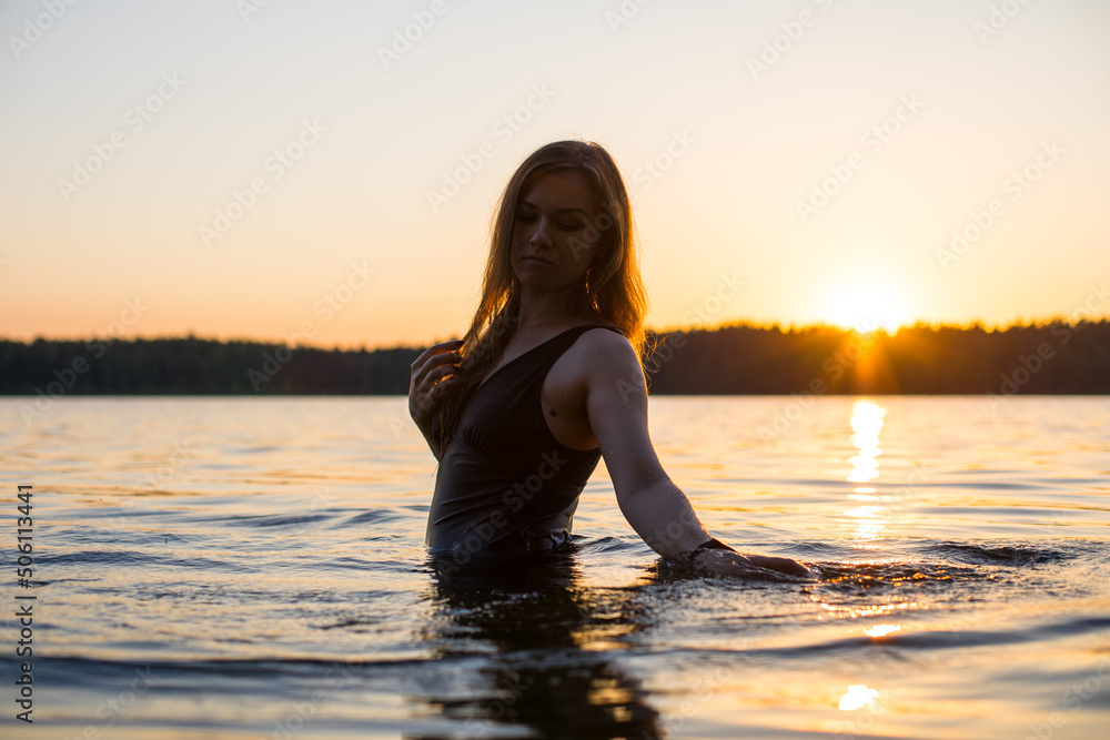 Beautiful Russian girl in a long black swimsuit swims outside the city on the lake in the rays of sunset or dawn. Splashing water on the background of the sun. Happy girl. Beautiful Russian girl in a long black swimsuit swims outside the city on the lake in the rays of sunset or dawn. Splashing water on the background of the sun. Happy girl.