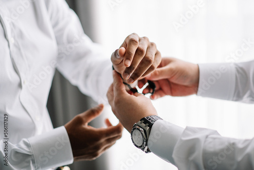 African American groom and his friends prepare for the wedding. Grooms man helps the groom to put on black cufflinks on white sleeve of the shirt. Details of the morning of the wedding day.