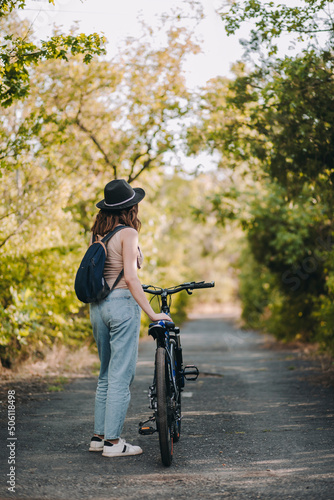 Obraz na płótnie Back view of young woman in a hat posing for camera with bike in a summer peaceful park. Healthy lifestyle and recreation w salonie