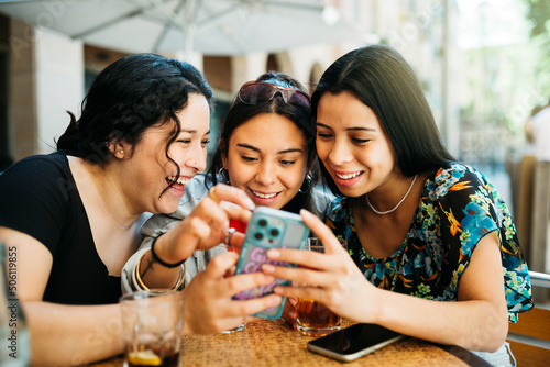 Fotografie Three young women looking at a phone on terrace of a restaurant