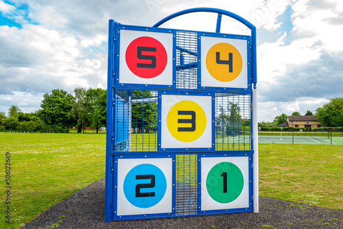 Newly installed colourful playground equipment seen on a large public field within a village community. The object is to hit a ball at the number targets.