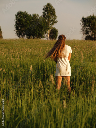 Young woman with long hair in a white dress stands with in the summer in a field. Sunny day, rear view. Summer inspiration. Juicy green grass and a beautiful slender girl. Trees in the background.