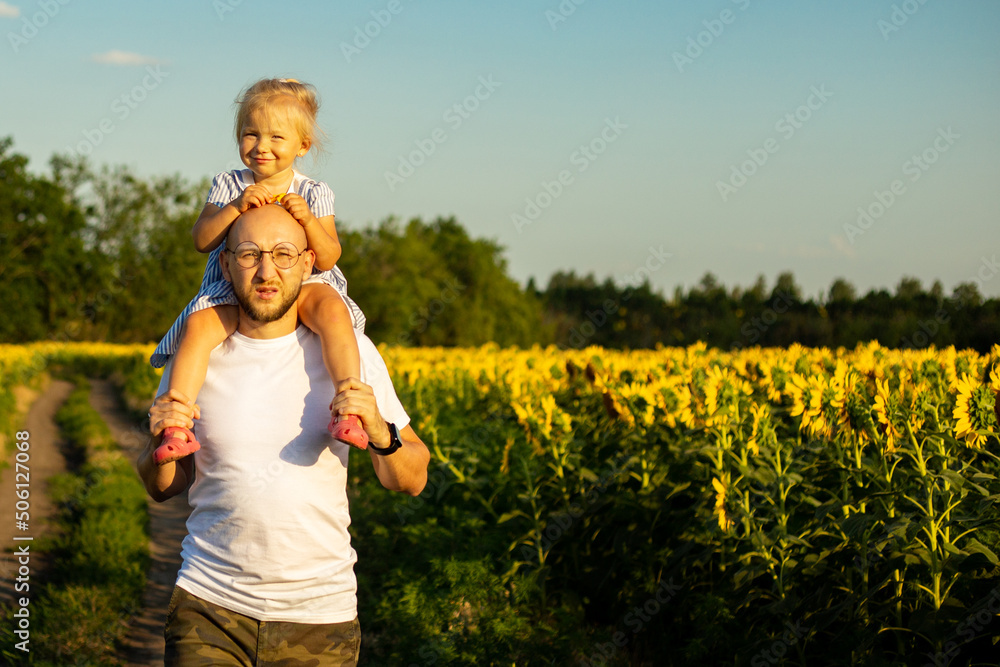 Young man wearing glasses carries a child on his shoulders across a sunflower field