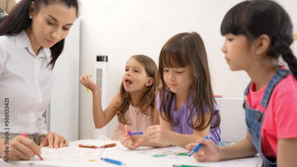 Woman teacher teaching little girl to paint color book on the table in classroom,kindergarten education school.Multi-ethnic preschool teacher and students in classroom.Kindergarten and study concept. Woman teacher teaching little girl to paint color book on the table in classroom,kindergarten education school.Multi-ethnic preschool teacher and students in classroom.Kindergarten and study concept.