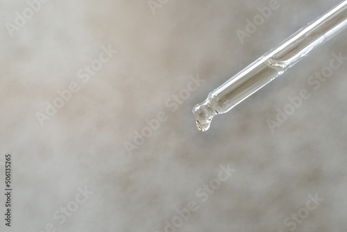Transparent glass pipette with essential oil or serum on a light gray concrete background. A drop of cosmetic oil falls. Health care. Copy space.
