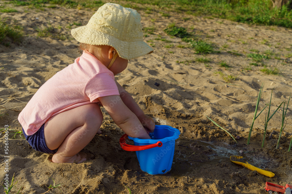 little baby playing with a blue plastic bucket filled with water on a background of sand, a child with bare feet pours water from a bucket learns something new fun childhood in the summer on vacatio little baby playing with a blue plastic bucket filled with water on a background of sand, a child with bare feet pours water from a bucket learns something new fun childhood in the summer on vacatio