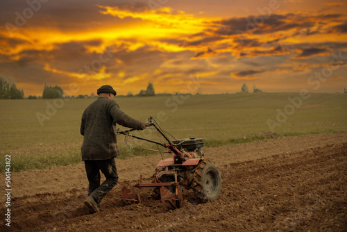 male farmer plows the soil of the garden with a walk-behind tractor cultivator at sunset.