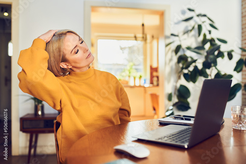Mature woman stretching neck while sitting at dinning table with laptop working from home