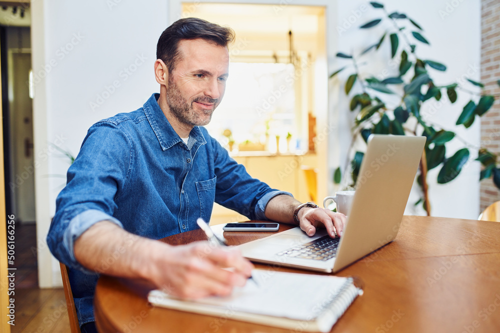 Mid adult man using laptop writing notes while at table working remotely Mid adult man using laptop writing notes while at table working remotely