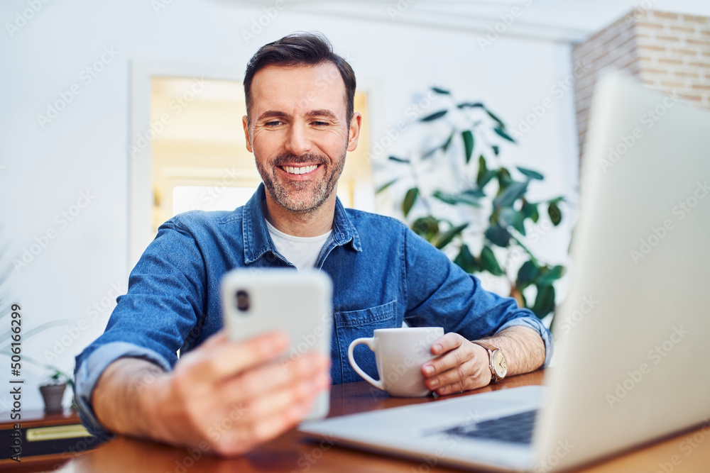 Portrait of happy relaxed man using phone and laptop while having coffee sitting at table at home Portrait of happy relaxed man using phone and laptop while having coffee sitting at table at home