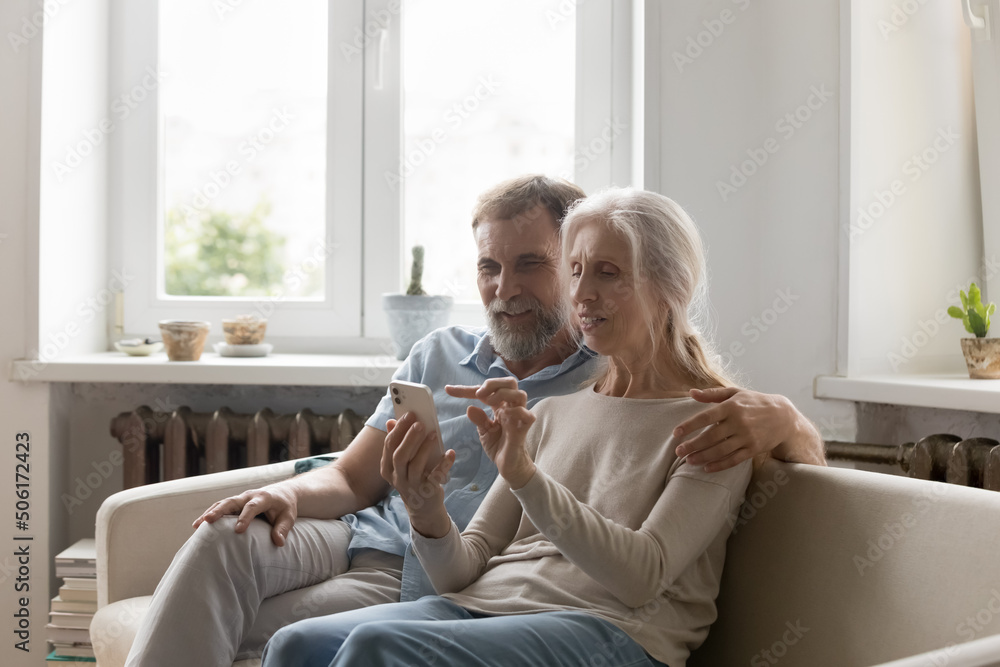 Positive focused senior couple using online app on smartphone, internet virtual payment service on mobile phone, making video call, shopping on we stores, resting on couch at home Positive focused senior couple using online app on smartphone, internet virtual payment service on mobile phone, making video call, shopping on we stores, resting on couch at home