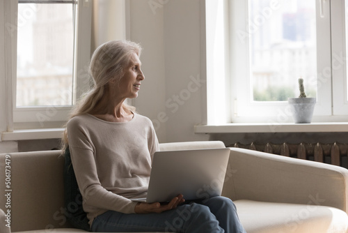 Happy pensive elder 70s woman holding laptop, resting on couch, looking at window away, thinking over webinar, workshop, smiling at good thoughts. Senior lady studying online, working from home